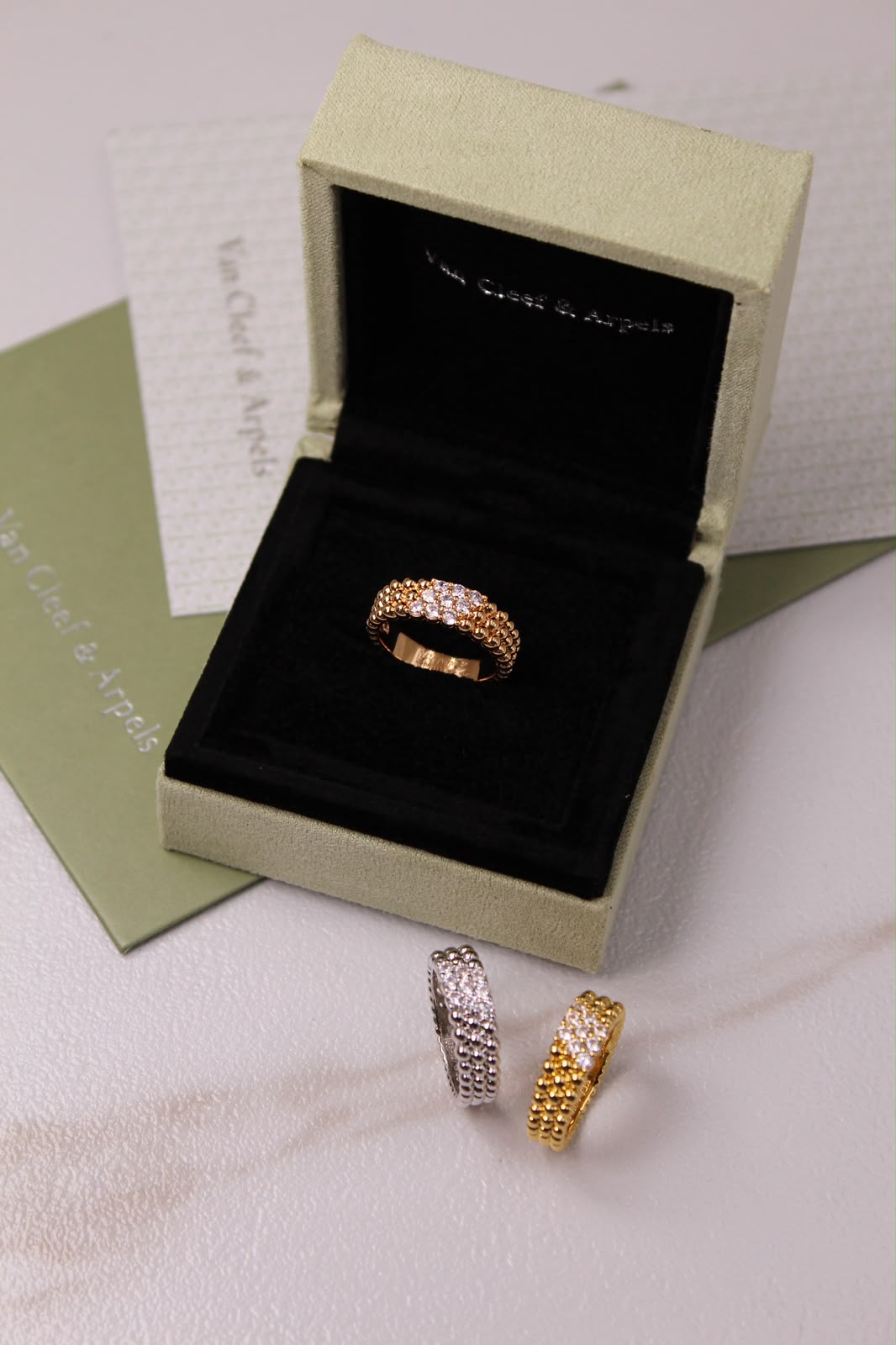 A gold eternity ring with pavé-set clear stones sits in an open light green jewelry box with a black velvet interior. Two additional rings, one gold and one silver, are placed in front on a light surface.